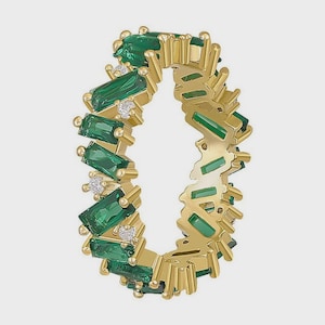 Könnte beinhalten: Ein goldfarbener Ring mit rechteckigen, smaragdgrünen Steinen und kleinen, klaren Akzentsteinen. Der Ring hat ein Zickzack-Design mit den grünen Steinen, die in einem sich wiederholenden Muster um das Band angeordnet sind. Dies ist ein Schmuckstück.