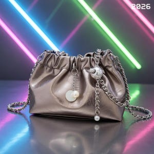 Puede incluir: Bolso plateado metálico con cordón y correa de cadena. El bolso tiene una cadena plateada y un dije de perla. El número "2026" está en la esquina superior derecha. Fondo con luces de neón.