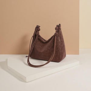 Könnte beinhalten: Eine braune Wildleder-Umhängetasche mit geknoteten Griffen. Die Tasche hat eine weiche, lockere Form und einen Kordelzugverschluss. Die Tasche steht auf einer weißen Oberfläche.