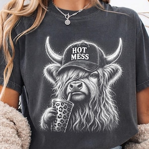 May include: A charcoal gray t-shirt featuring a white graphic of a Highland cow wearing a baseball cap with the text HOT MESS printed on it. The cow is holding a cup with a leopard print pattern. The shirt has a crew neckline and short sleeves.