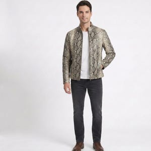 May include: A full-length shot of a person wearing a snakeskin-patterned jacket, a white t-shirt, dark gray jeans, and brown leather shoes. The jacket has a zipper closure and a stand-up collar. The background is plain white.