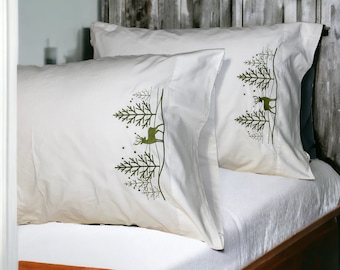 Puede incluir: Dos fundas de almohada blancas con árboles verdes bordados y un diseño de ciervo. Las fundas de almohada están sobre una cama blanca con una cabecera de madera.