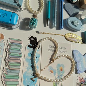 Pu&ograve; includere: Un assortimento di articoli di cancelleria e gioielli su una superficie bianca. Gli articoli includono una collana di perle, un braccialetto, penne, un segnalibro con un design a gatto e un piccolo barattolo pieno di pietre turchesi. &Egrave; visibile anche una carta con il testo "CULTIVATED OPIUM POPPY".