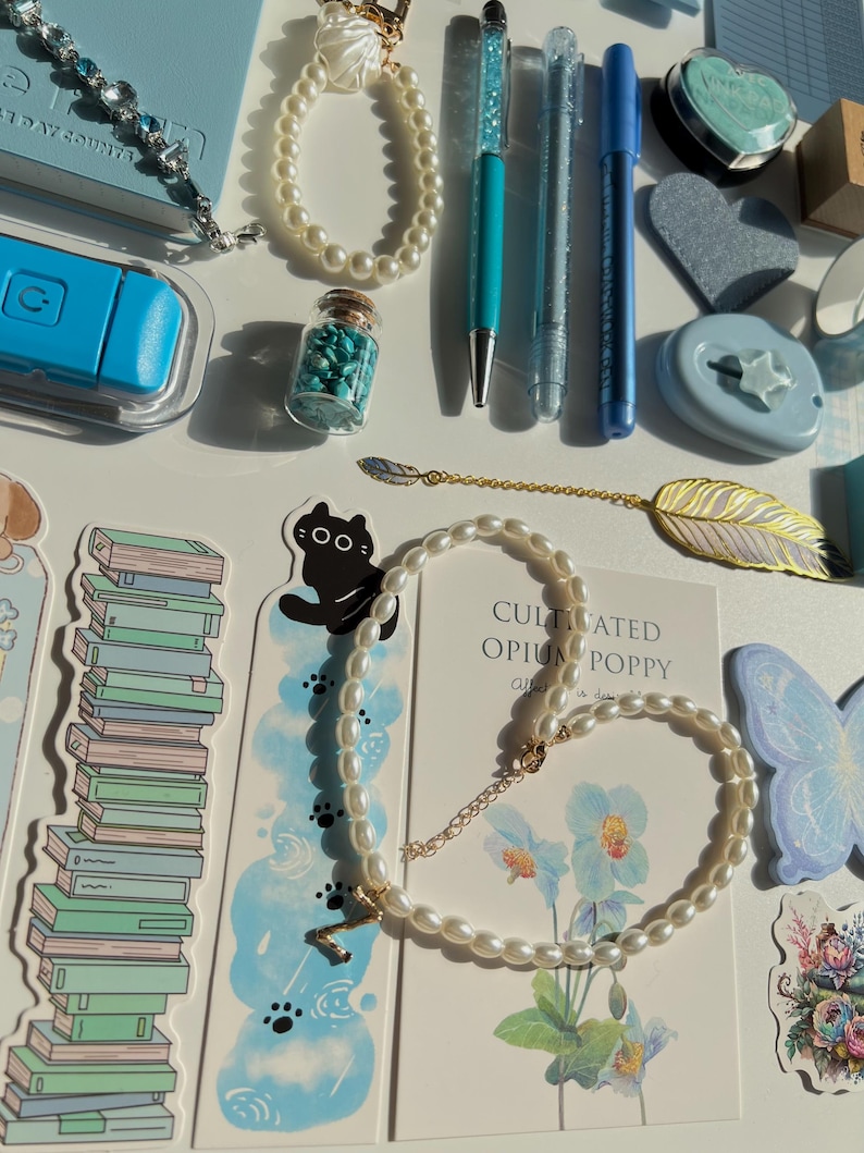 Pu&ograve; includere: Un assortimento di articoli di cancelleria e gioielli su una superficie bianca. Gli articoli includono una collana di perle, un braccialetto, penne, un segnalibro con un design a gatto e un piccolo barattolo pieno di pietre turchesi. &Egrave; visibile anche una carta con il testo "CULTIVATED OPIUM POPPY".