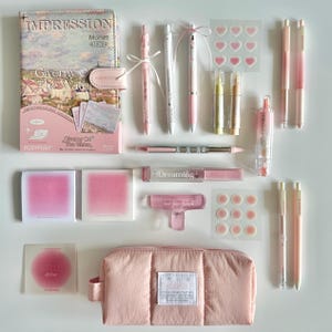 Puede incluir: Una colección de artículos de papelería rosas y blancos, que incluyen bolígrafos, pegatinas, un cuaderno y un estuche. El cuaderno presenta una pintura de Monet. El estuche es de tela acolchada rosa claro.