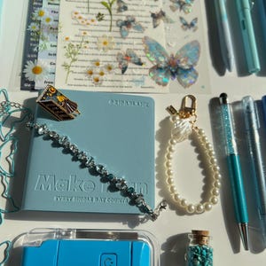 Pu&ograve; includere: Una collezione di cancelleria e accessori nei toni del blu e del bianco. Include un taccuino blu con la scritta "Make it happen", adesivi a farfalla, penne, un braccialetto, un portachiavi di perle e una piccola bottiglia di pietre turchesi.