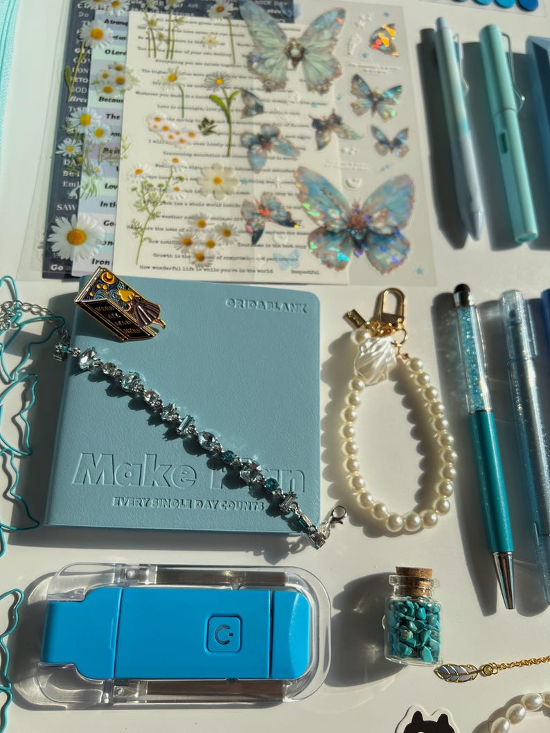 Pu&ograve; includere: Una collezione di cancelleria e accessori nei toni del blu e del bianco. Include un taccuino blu con la scritta "Make it happen", adesivi a farfalla, penne, un braccialetto, un portachiavi di perle e una piccola bottiglia di pietre turchesi.
