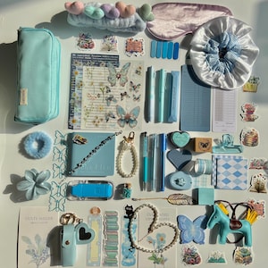 Op de afbeelding: Een verzameling blauwe en pastelkleurige briefpapier en accessoires, waaronder een etui, pennen, stickers, een slaapmasker, een scrunchie en een notitieboek. Decoratieve items zijn vlinder- en bloemmotieven, een paardenbeeldje en een parelketting.