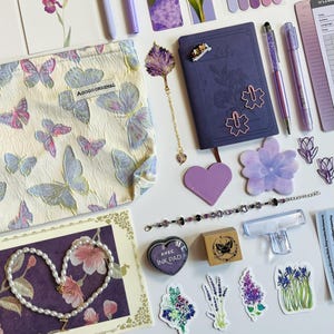 Puede incluir: Un surtido de artículos de papelería y accesorios morados y lavanda, que incluyen una bolsa con estampado de mariposas, un diario, bolígrafos, pegatinas y un collar de perlas. Los artículos están dispuestos sobre una superficie blanca, creando una colección visualmente atractiva.