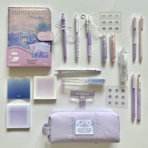 Peut inclure: Divers articles de papeterie lavande, dont des stylos, une trousse, des notes autocollantes et des autocollants. Un carnet avec un design "Waterloo Bridge" est également présent. Les articles sont disposés sur une surface blanche.