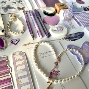 May include: An assortment of purple stationery and accessories, including pens, a heart-shaped patch, washi tape, and a pearl necklace with a gold initial pendant. Decorative butterfly and floral stickers are also visible.