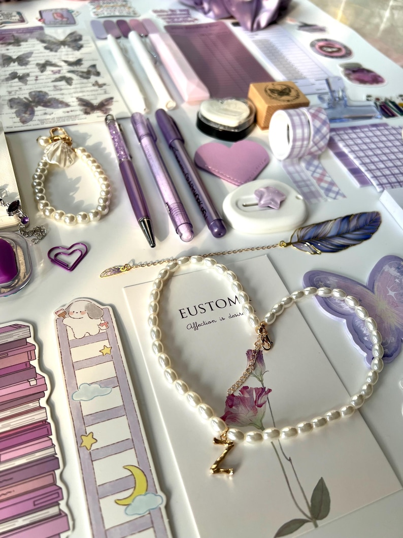 May include: An assortment of purple stationery and accessories, including pens, a heart-shaped patch, washi tape, and a pearl necklace with a gold initial pendant. Decorative butterfly and floral stickers are also visible.