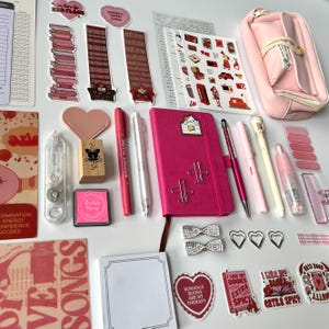 Puede incluir: Una colección de artículos de papelería rosas y rojos, que incluyen un diario, bolígrafos, pegatinas y clips decorativos. Los artículos están dispuestos sobre una superficie blanca. El diario tiene clips plateados con forma de avión.