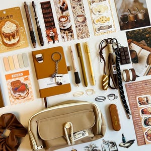 Puede incluir: Un surtido de artículos de papelería, incluyendo bolígrafos, marcapáginas, pegatinas, un cuaderno y una bolsa pequeña, todo en una paleta de colores cálidos y neutros. Los artículos tienen un tema de café, con ilustraciones de tazas de café y postres.