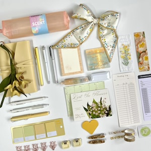 Puede incluir: Un conjunto de papelería que incluye un estuche color melocotón, un lazo floral, un cuaderno amarillo con dijes de flores, varios bolígrafos, notas adhesivas, marcapáginas, un registro de lectura, un rastreador de libros y clips.