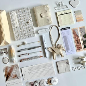 Puede incluir: Una colección de artículos de papelería que incluye un cuaderno de tweed crema, una cartera de color crema con textura, un bloc de notas, un registro de lectura, un dije de pegaso, bolígrafos, marcapáginas, clips de corazón y varias pegatinas.