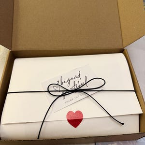 May include: A white package tied with black string and a bow, inside a cardboard box. A small card with the words "beyond grateful" and a red heart sticker are attached to the package.