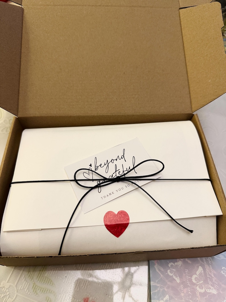 May include: A white package tied with black string and a bow, inside a cardboard box. A small card with the words "beyond grateful" and a red heart sticker are attached to the package.