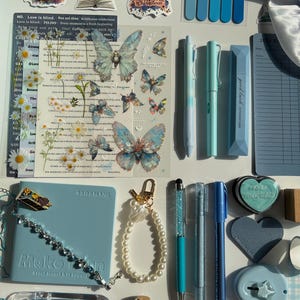 Pu&ograve; includere: Una collezione di articoli di cancelleria e accessori nei toni del blu e del bianco. Include penne, adesivi, un diario, un braccialetto, un portachiavi e una piccola bottiglia con pietre turchesi. Sono presenti anche adesivi decorativi a farfalla.