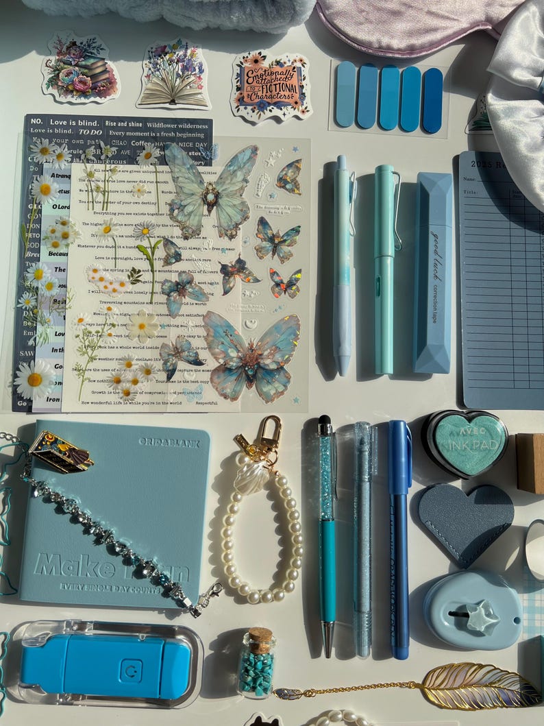 Pu&ograve; includere: Una collezione di articoli di cancelleria e accessori nei toni del blu e del bianco. Include penne, adesivi, un diario, un braccialetto, un portachiavi e una piccola bottiglia con pietre turchesi. Sono presenti anche adesivi decorativi a farfalla.