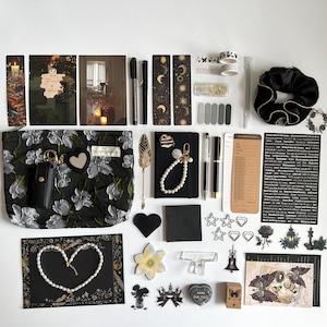 May include: An assortment of stationery and accessories, including a black floral pouch, notebooks, pens, stickers, and washi tape. The items are arranged on a white surface, with a dark, gothic aesthetic. There are also decorative elements like a pearl necklace and a black scrunchie.