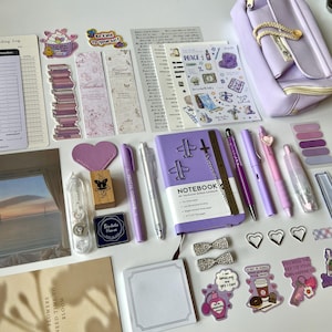 Op de afbeelding: Een verzameling paarse en roze briefpapierartikelen, waaronder pennen, een notitieboek, stickers en een etui. Het notitieboek heeft een zilveren ketting en vliegtuigvormige paperclips. Decoratieve hartvormige paperclips en stickers zijn ook aanwezig.