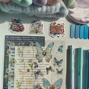 Pu&ograve; includere: Una collezione di cancelleria e accessori, tra cui una fascia blu con dettagli a forma di cuore, adesivi con disegni floreali e a farfalla e penne blu. Gli adesivi presentano testo come "Emotionally Attached to Fictional Characters."