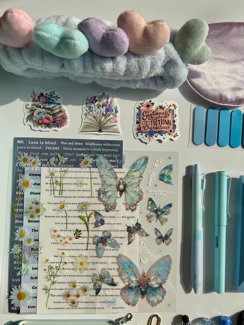 Pu&ograve; includere: Una collezione di cancelleria e accessori, tra cui una fascia blu con dettagli a forma di cuore, adesivi con disegni floreali e a farfalla e penne blu. Gli adesivi presentano testo come "Emotionally Attached to Fictional Characters."