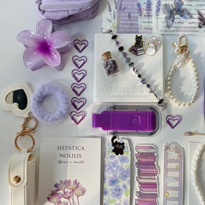 May include: A collection of purple and white stationery items, including a small bag, flower-shaped clip, heart-shaped paper clips, a bracelet, a notebook, and stickers. The notebook reads "Make Plans Every Single Day Counts".