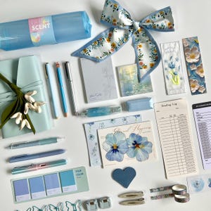 May include: A collection of stationery items including a light blue pencil case, a floral ribbon, pens, a reading log, bookmarks, sticky notes, and decorative paper clips. The set features various shades of blue, white, and floral patterns.