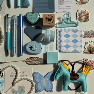 Pu&ograve; includere: Un assortimento di articoli di cancelleria in tonalit&agrave; di blu, tra cui penne, adesivi, un timbro e nastro decorativo. &Egrave; presente anche una piccola statuina di cavallo azzurro con sella marrone, insieme a una collana di perle e un adesivo a farfalla.