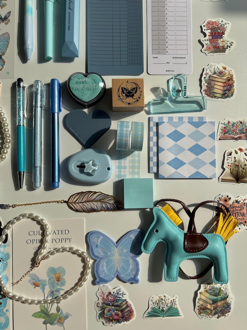 Pu&ograve; includere: Un assortimento di articoli di cancelleria in tonalit&agrave; di blu, tra cui penne, adesivi, un timbro e nastro decorativo. &Egrave; presente anche una piccola statuina di cavallo azzurro con sella marrone, insieme a una collana di perle e un adesivo a farfalla.