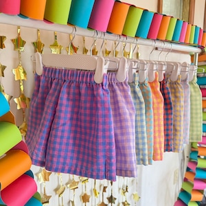 May include: A row of multiple elastic-waist skirts hanging on white plastic hangers. Each skirt features a different colored gingham check pattern, including blue and pink, purple, light blue, yellow, orange, and green variations.