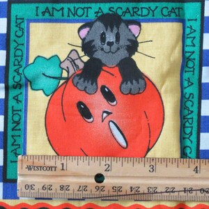 May include: A Halloween-themed fabric square featuring a cartoon cat perched on a carved orange pumpkin. The text "I AM NOT A SCARDY CAT" is printed around the image. The border includes blue and white checkered patterns and a black background.
