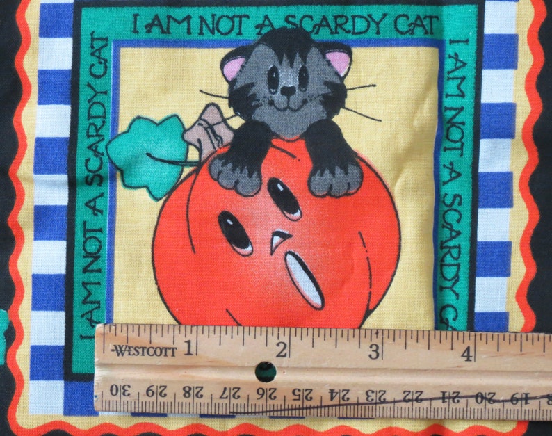 May include: A Halloween-themed fabric square featuring a cartoon cat perched on a carved orange pumpkin. The text "I AM NOT A SCARDY CAT" is printed around the image. The border includes blue and white checkered patterns and a black background.