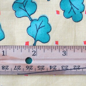 May include: Close-up of a fabric with a light yellow background. The fabric features a repeating pattern of teal green leaves, small red squares, and cartoon figures. A wooden ruler is placed across the fabric, showing measurements in inches.