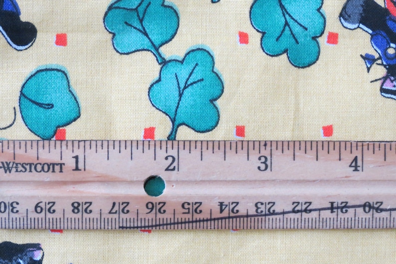 May include: Close-up of a fabric with a light yellow background. The fabric features a repeating pattern of teal green leaves, small red squares, and cartoon figures. A wooden ruler is placed across the fabric, showing measurements in inches.