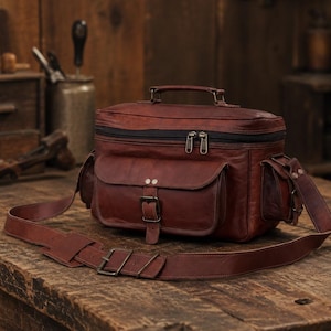 Pode incluir: Uma bolsa de câmara em couro castanho escuro com uma pega superior, um compartimento principal com fecho de correr e um bolso frontal fixado com uma fivela. A bolsa inclui bolsos laterais e uma alça ajustável com ferragens metálicas.