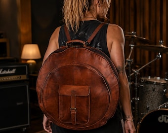 May include: A large, circular brown leather backpack featuring a prominent front pocket with a vertical strap and metal buckle closure. The bag includes two shoulder straps and a top handle, with visible stitching along the edges of the leather.