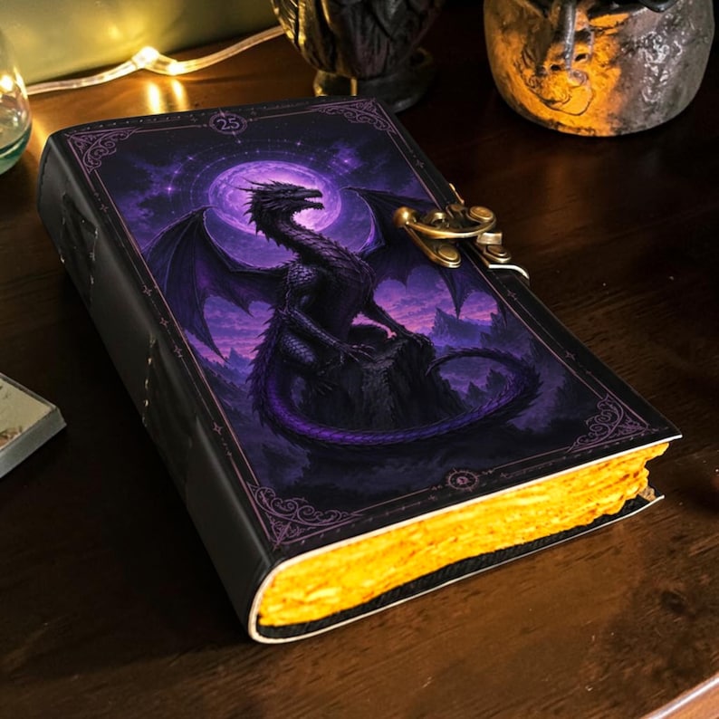 May include: A rectangular journal with a black cover featuring a purple dragon illustration against a full moon and mountain landscape. The book has a metallic gold-toned latch closure and thick, textured pages with a vibrant yellow-gold edge finish.