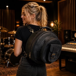 May include: A round black leather backpack featuring a front pocket with a flap and a buckle closure. The bag includes a top handle, adjustable shoulder straps with gold-toned metal hardware, and a zippered main compartment for storage.