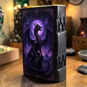 May include: A rectangular black leather-bound journal featuring a purple-toned illustration of a dragon perched on a rock under a full moon. The spine is secured with black cord lacing in a crisscross pattern, and the number 25 is printed at the top.