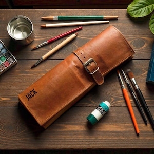 May include: A tan leather artist's brush roll with the name "JACK" printed on the front. The roll is secured with a buckle and contains various paint brushes and pencils. A tube of paint and a tin of water are also visible.