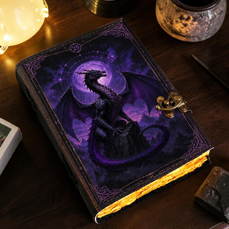 May include: A rectangular journal featuring a dark purple cover with a dragon perched on a rock under a full moon. The cover includes ornate corner borders, a circular emblem with the number 25, and a metallic brass-colored latch closure.