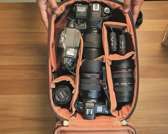 May include: A brown camera backpack with an open interior featuring orange lining and padded dividers. The bag contains two digital cameras, three camera lenses, a flash unit, and two small electronic accessories organized in separate compartments.