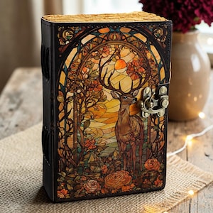 Pode incluir: Um diário retangular encadernado em couro com uma estampa estilo vitral de um veado em uma floresta com flores laranja e um pôr do sol. A capa é presa com uma trava metálica e possui bordas pretas com detalhes de costura visíveis.