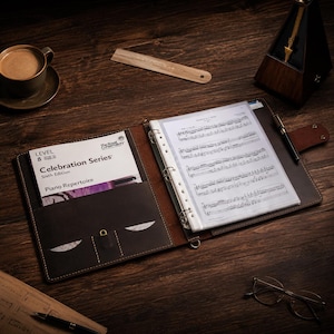 Può includere: Un raccoglitore in pelle marrone con spartiti e un libro intitolato "Celebration Series Piano Repertoire" su una scrivania di legno. Una tazza di caffè, un metronomo e una penna sono anche sulla scrivania.