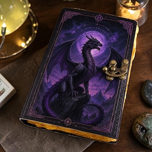 May include: A rectangular journal with a dark cover featuring a purple dragon perched on a rock against a full moon background. The cover has a decorative metal latch on the right side and the number 25 printed in a small circle at the top center.