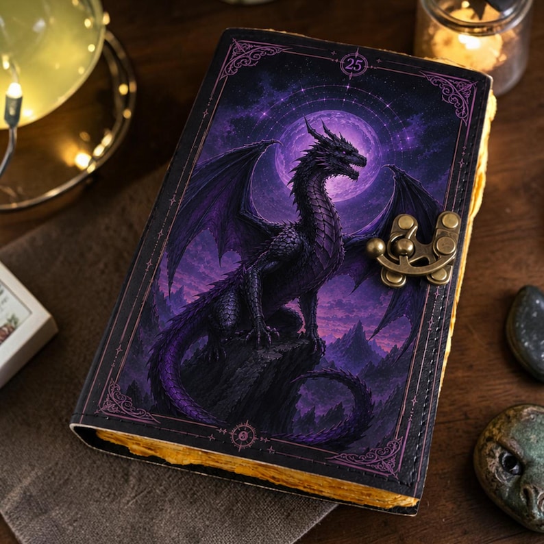 May include: A rectangular journal with a dark cover featuring a purple dragon perched on a rock against a full moon background. The cover has a decorative metal latch on the right side and the number 25 printed in a small circle at the top center.