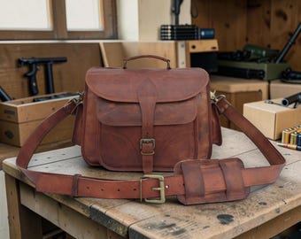 May include: A brown leather messenger bag with a top handle, flap closure, and adjustable shoulder strap. The bag features a buckle closure and a padded shoulder rest. The bag measures approximately 12 inches in height and 14 inches in width.